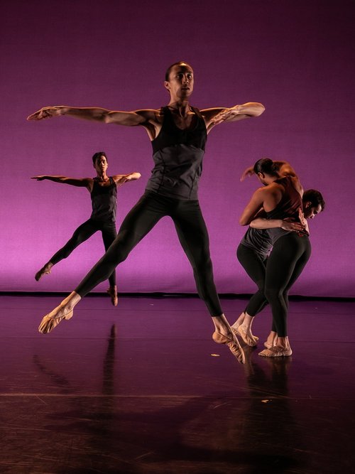 two maile dancers in tight grey/black outfits are suspended in air in a pose where there legs are extended wide in a inverted v jump. there arms are angular, one extended to the side the other bent two dancers huddle to their side 